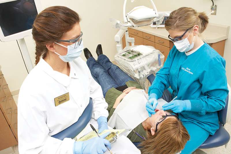 Two dentists stand over a patient inspecting her teeth with dentist tools