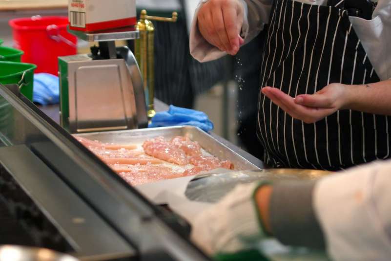 A student dropping a pinch of large, grainy salt over raw chicken breasts on a baking sheet.