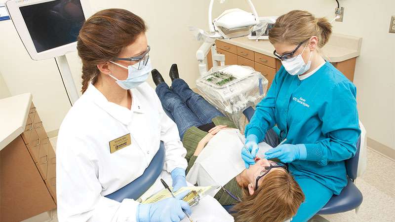 Two dentists stand over a patient inspecting her teeth with dentist tools