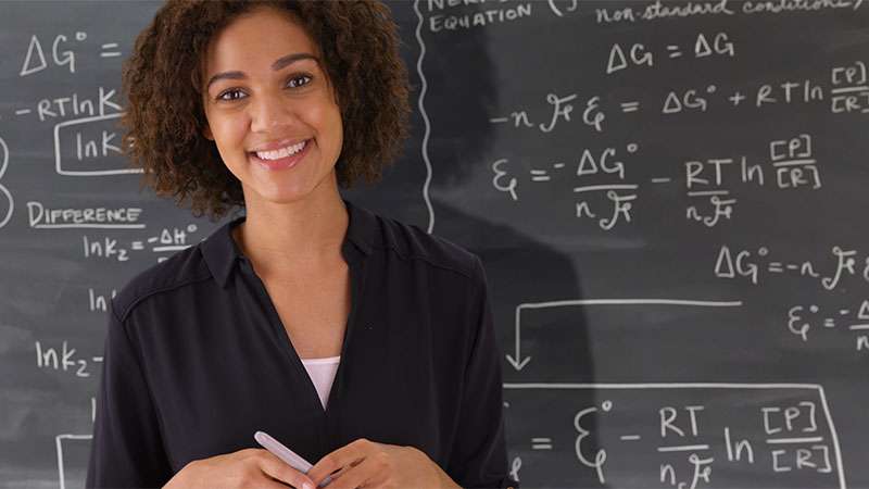 A woman smiles standing in front of a chalkboard full of math equations