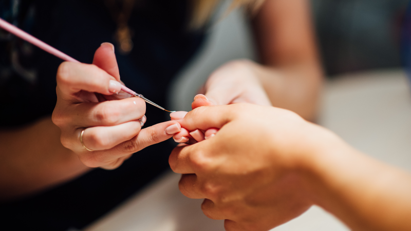 Using a brush, a Nail Technologist applies a gel finish to a client’s nails on her left hand.