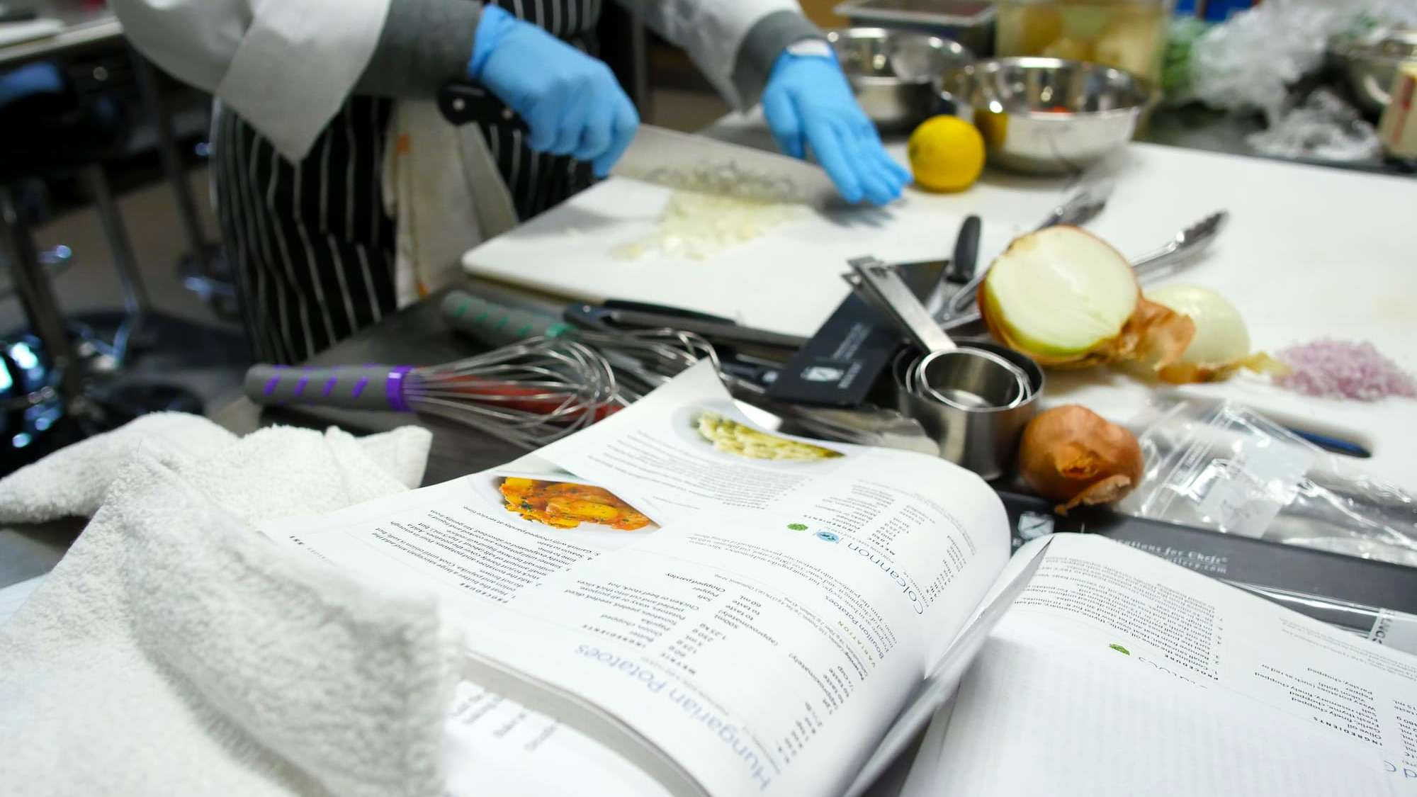 Close-up view of a busy kitchen work space. A student dices an onion as other tools and ingredients are kept nearby.