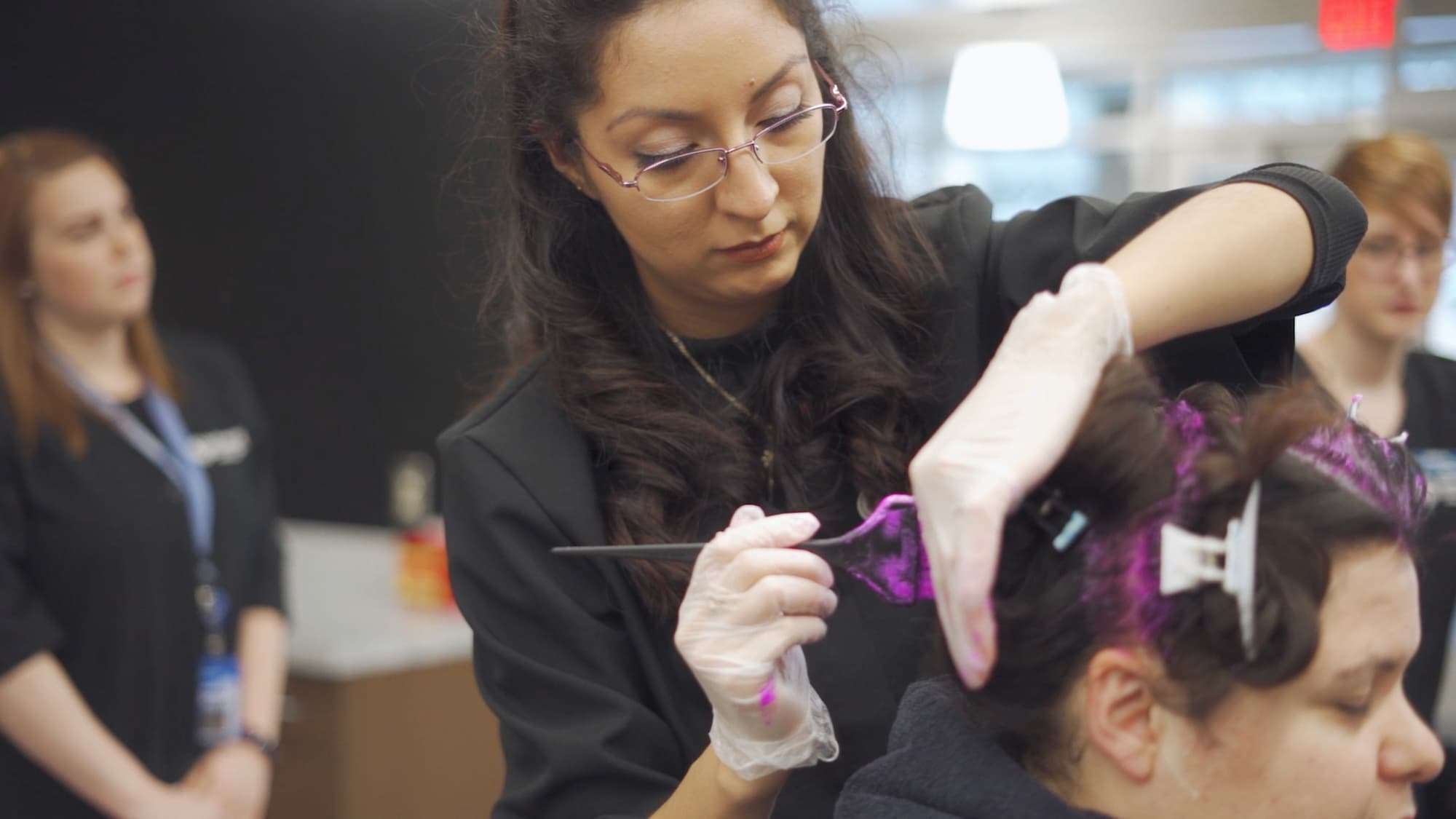 A hair stylist wears disposable gloves as she brushes purple hair dye into her client's hair.