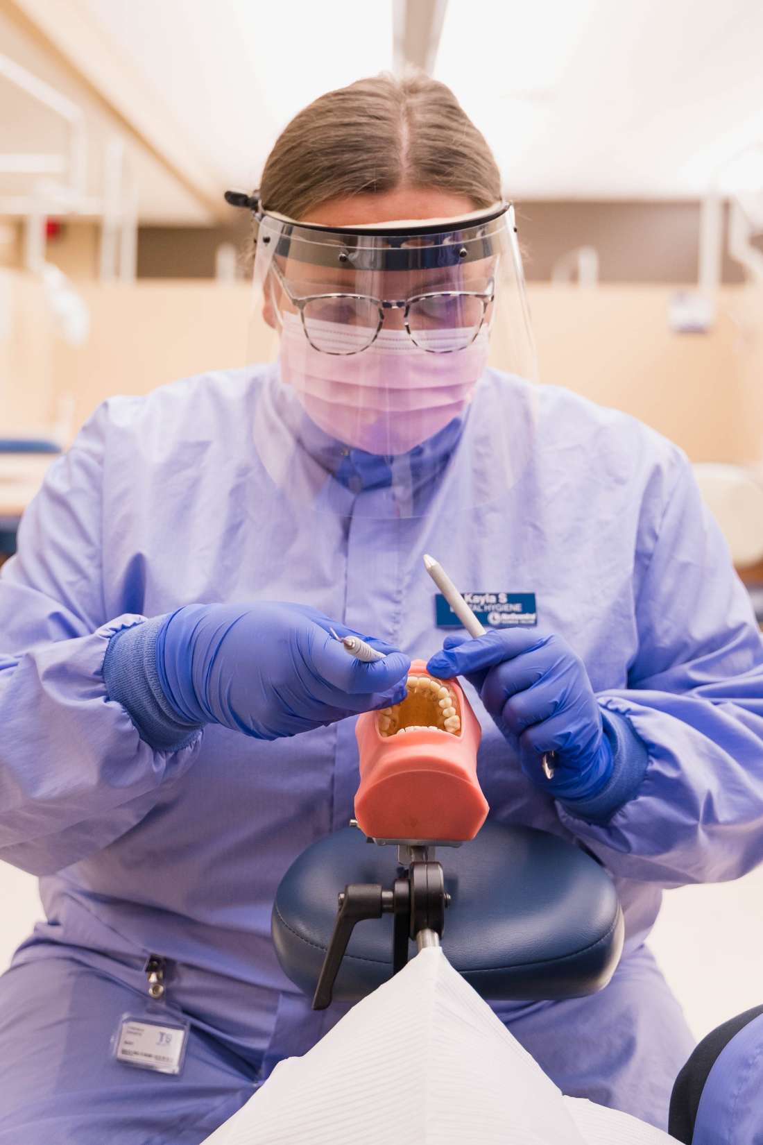 A dental hygienist dressed in medical garbs with a face shield performs a medical procedure on dental mouth model.