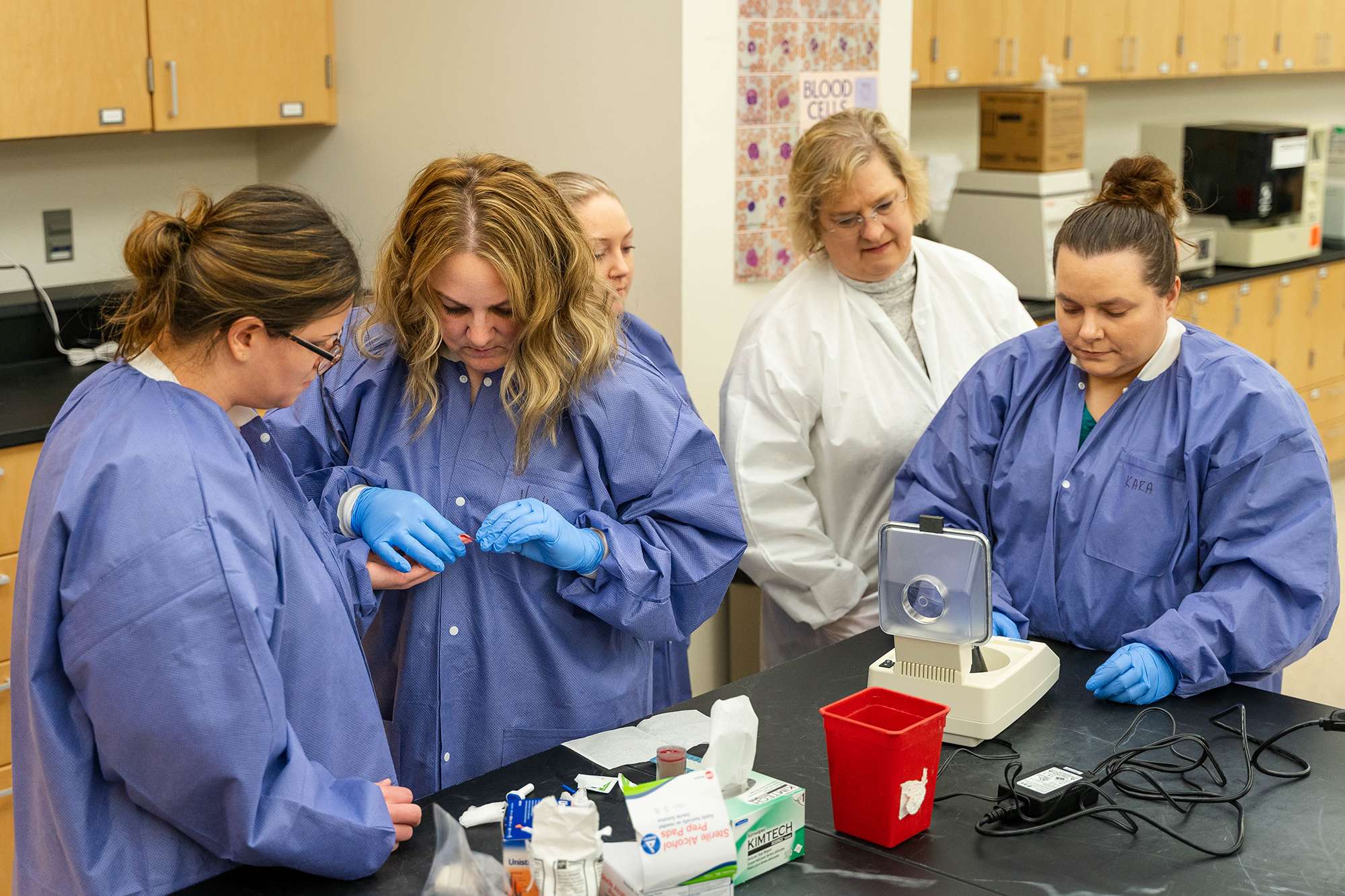 Four Medical Assistants and an instructor are working on blood samples together inside of a medical lab.