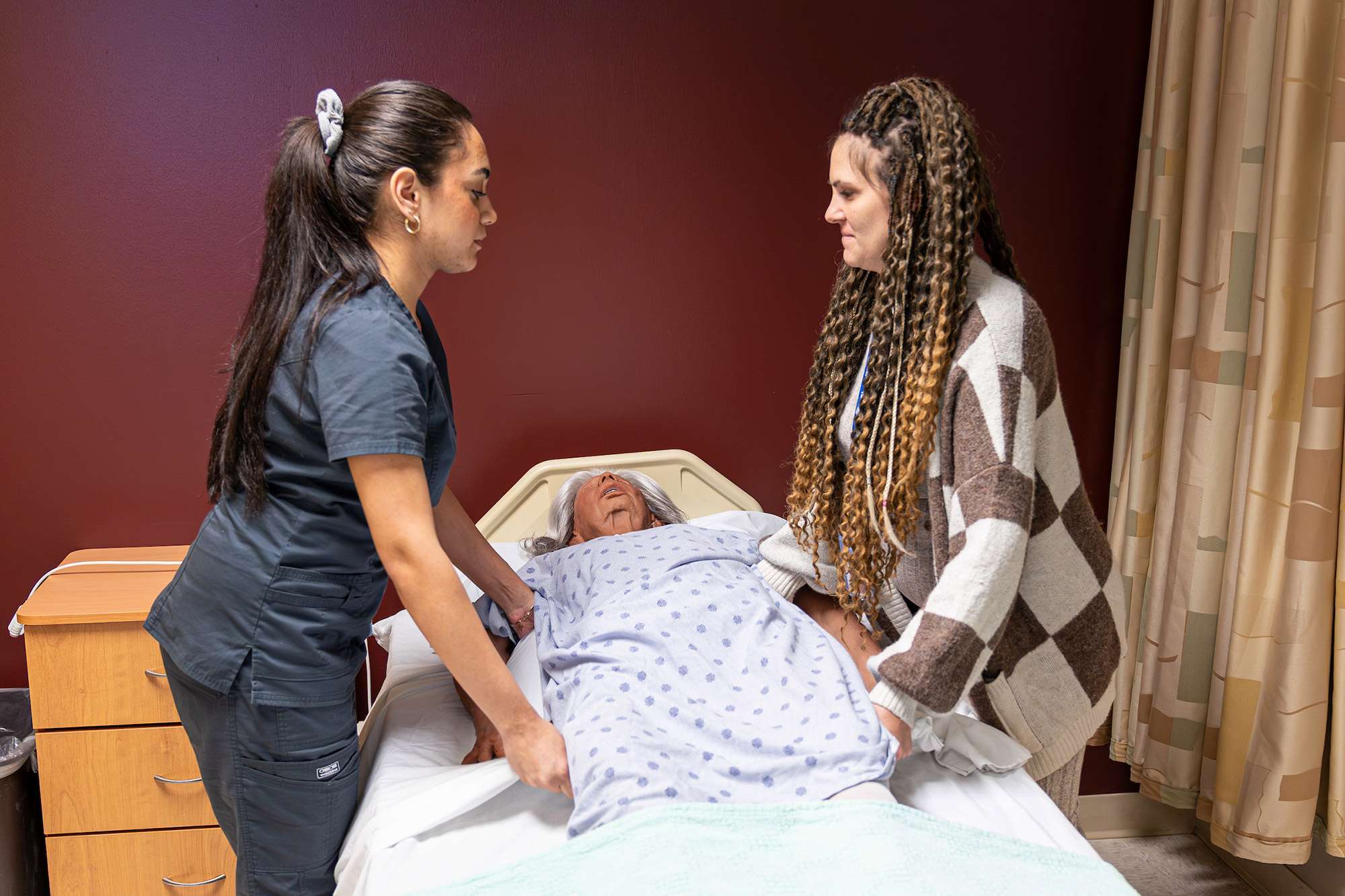 Two certified nursing assistants help each other move a mannequin of an elderly patient.