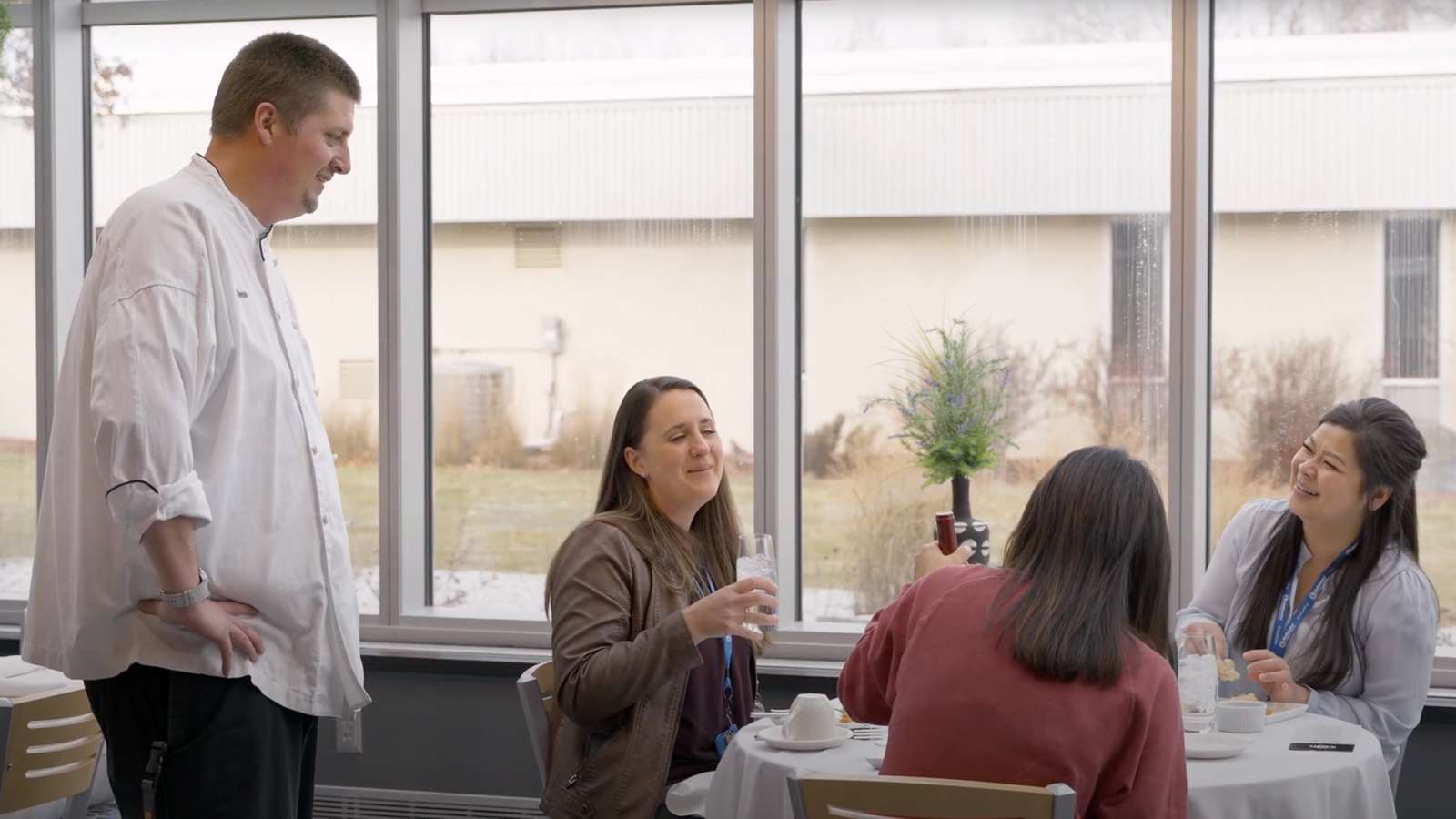 A group of NTC employees having lunch while seated at a table in Spoons Restaurant while a Culinary instructor stands nearby.