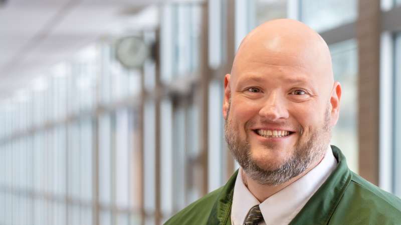 Shawn Sullivan smiling while standing in a hallway with a wall of windows behind him.