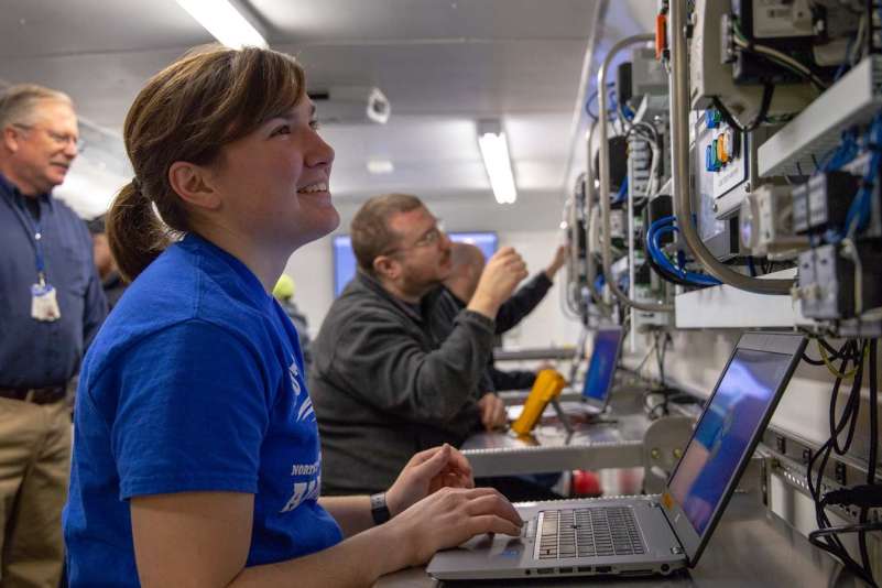 A student sits at a table in the Emerging Technologies Mobile Lab, as she works on a laptop and looks at controls mounted on the wall.