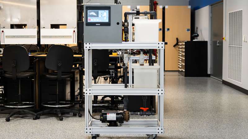 A process control training station on wheels, featuring a touchscreen interface, clear fluid reservoirs, pipes, and a pump system. The unit is framed in aluminum, set in a lab with workstations and storage cabinets in the background.